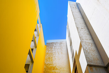Two buildings rise side by side, one yellow, the other weathered white, framing a sky above....