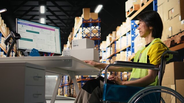 Asian man wheelchair user typing tracking numbers on a computer software, updating parcel status in real time. Staff managing order fulfillment in a logistics center to sort inventory. Camera A.