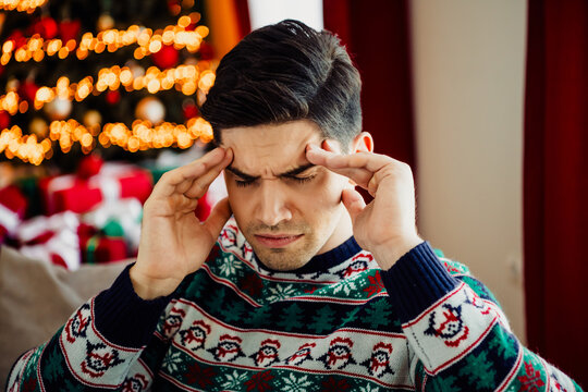 Young man in festive Christmas sweater experiencing holiday fatigue with gifts and tree in cozy illuminated home