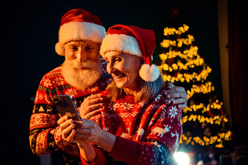 Warm elderly couple in matching santa hats enjoy a playful moment as they view a smartphone beside a glowing christmas tree and festive lights in a cozy home setting