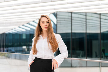 Confident young businesswoman looking at camera. Professional lady with long brown hair in businesswear standing outside office building in natural light