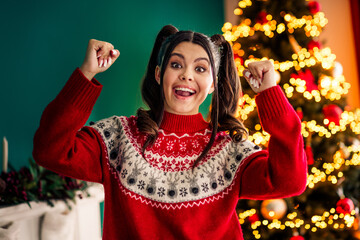 Excited young woman wearing a festive holiday sweater enjoying the spirit of Christmas at home by...