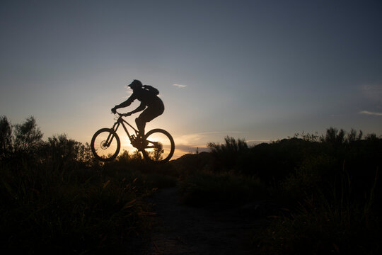 Fotografia de silueta de una mujer saltando en una bici de monta&ntilde;a al atardecer, donde su silueta sobre la bici resalta ante el sol y el cielo de verano