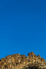 A summer landscape features a rocky stone wall beneath a blue sky and a passing airplane.