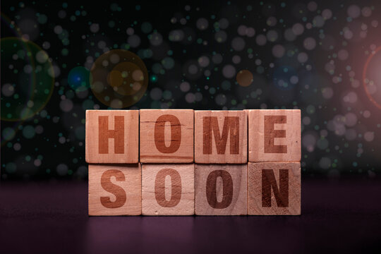 Wooden blocks spelling out the phrase HOME SOON on a dark background with bokeh lights, representing anticipation and return.