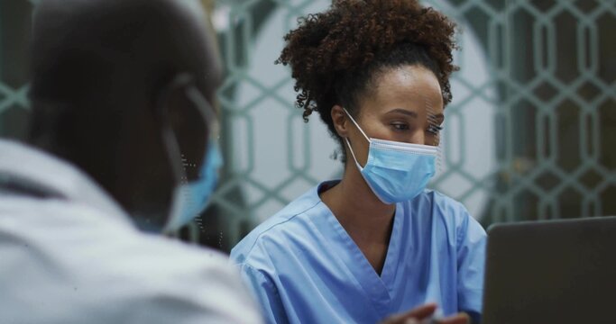 Nurse in blue scrubs and physician wearing masks reviewing records at nurses station, with laptop