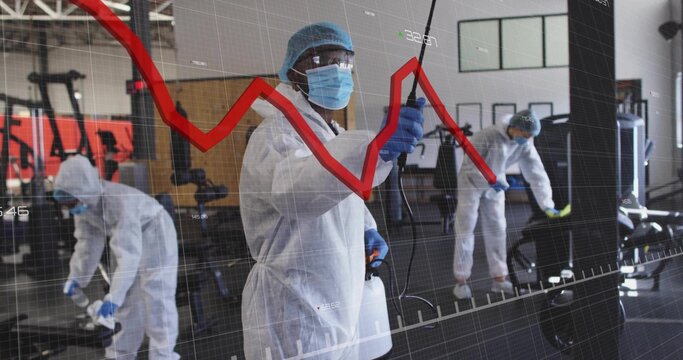 Disinfecting man in PPE using sprayer on machines in gym, with grid, red line graph overlay