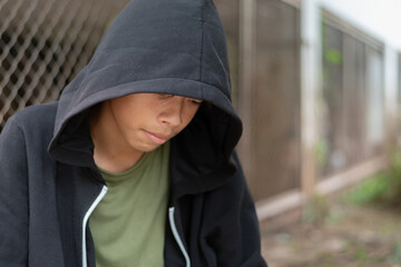 Young man in a black hooded sweatshirt looking down with a sad, pensive expression, suggesting feelings of depression, isolation, and inner struggle in an outdoor setting. © Sophon_Nawit