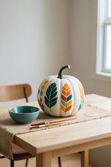 Photorealistic white pumpkin with colorful geometric leaf patterns on light wooden table, teal bowl, burlap, paintbrushes, soft window light, neutral background and chair
