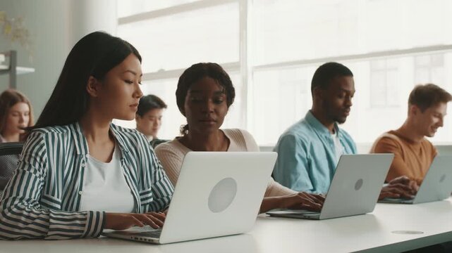 A group of diverse students is focused on their laptops during a study session at a university. They collaborate in a bright, modern classroom filled with natural light.