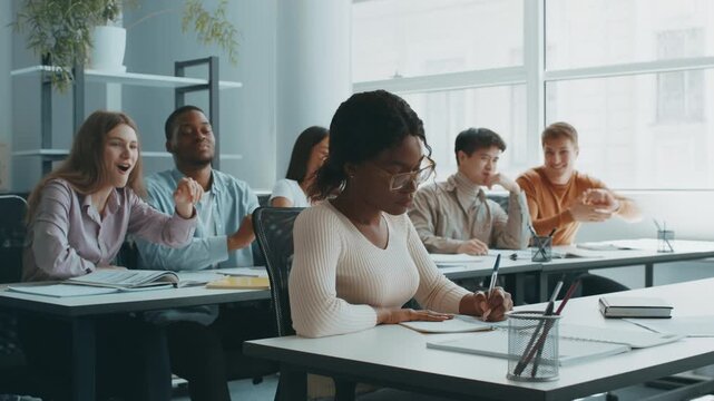 A group of diverse college students is working together in a bright, spacious classroom. One student focuses intently on taking notes while others bullying her - Powered by Adobe