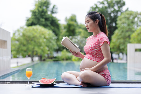 A pregnant woman in a red patterned outfit sits beside a pool reading a book, with watermelon and orange juice nearby. The scene expresses peacefulness, self-care, and healthy pregnancy lifestyle.