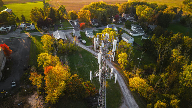 Communication tower near small suburban houses surrounded by autumn trees and green fields, captured from above in warm evening light.
