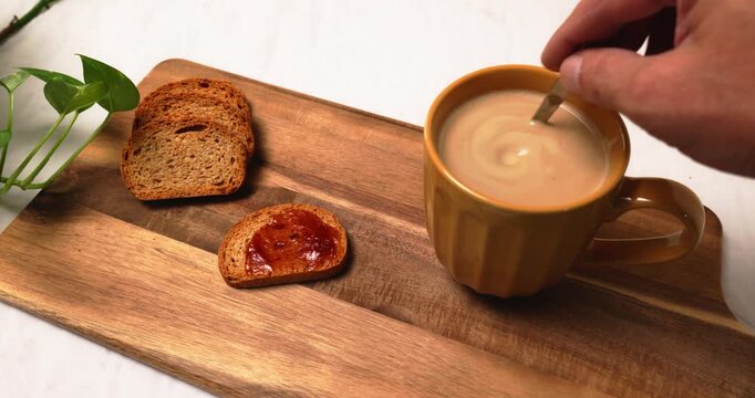 Hand stirring cup of coffee with milk located on wooden board with slices of toasted bread and jam for breakfast.