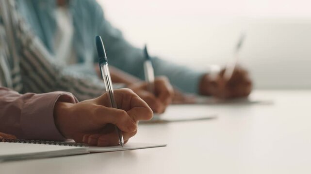 Diverse group of college students focus on their studies, writing notes with pens on notebooks. They are collaborating in a bright learning environment, emphasizing education and teamwork.