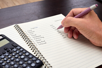 A person calculating and writing down a list of monthly household expenses.