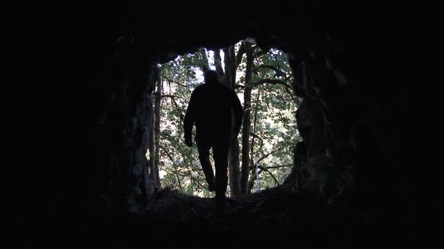Hiker walking into frame from dark cave that empties into sunshine filled forest.