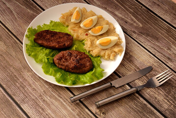 Two fried meat cutlets on a lettuce leaf and mashed potatoes with sliced boiled chicken egg on a wooden table.
