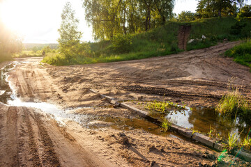 Rugged dirt path with watery ruts and glinting puddles under a bright sun. Tire tracks crisscross...