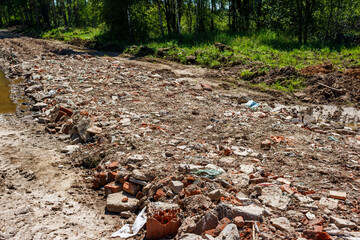 A chaotic landscape of construction debris and brick fragments lit by sun. A messy mix of rubble on an unkempt road near a green forest