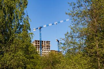 Tower cranes busy on a high-rise construction site, structure rising amidst green trees under a clear blue sky