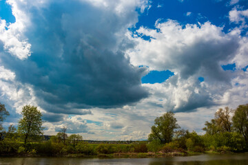 Majestic stormy clouds dominate, some fluffy white, others brooding grey, with vivid blue sky peeking through. Lush riverbanks and trees below