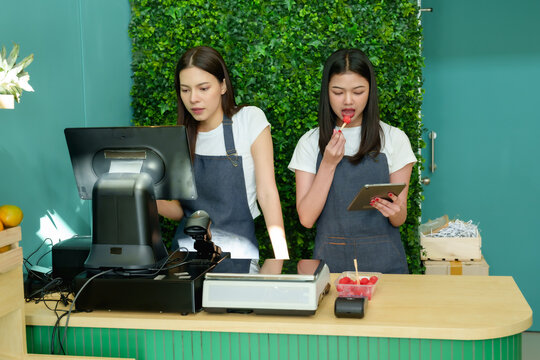 Two caucasian adult female fruit shop vendors wearing aprons using point of sale system together while one eats strawberry and another checks screen during fresh produce store setup - Powered by Adobe