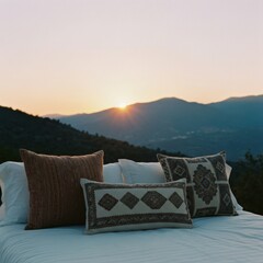 Three Decorative Pillows On A White Bed Against A Scenic Mountain Sunset Backdrop