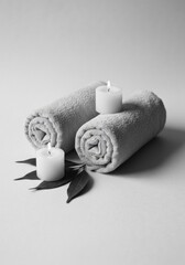 Two Rolled White Spa Towels With Lit White Candles And Green Leaves On A Light Gray Textured Background