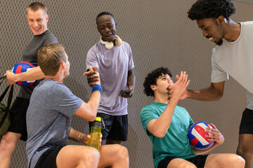 Diverse male volleyball partners resting in sports hall holding volleyballs and water bottle