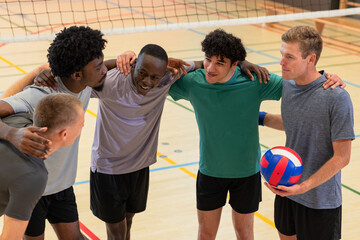 Obraz premium Diverse male partners huddling on gym volleyball court holding volleyball beneath net posts