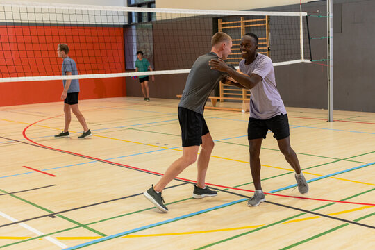 Diverse male partners exchanging high five at net inside gymnasium during volleyball match