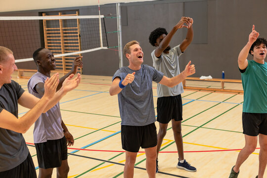 Diverse male volleyball team clapping, raising arms on gym court under net with water bottle