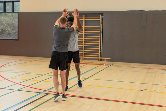 Diverse male workout partners raising arms overhead in gymnasium by climbing frame, court lines - Powered by Adobe
