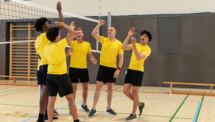 Diverse male teammates giving high-fives at volleyball net on gym court with wall bars