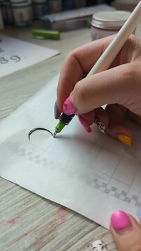 Close-up of person creating elegant calligraphy with pen on white paper, showing artistic handwriting and precision. Female hand with colorful manicure practicing calligraphy, drawing graceful lines.