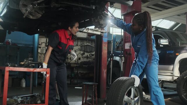Latin female mechanic worker fixes automotive wheels and talks to a Black coworker who maintains car suspensions in garage station, technician engineering service jobs in vehicle transport industry.