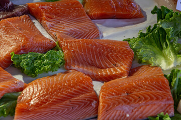 A close-up, high-angle view of multiple bright orange raw salmon fillets, with distinctive white fat lines, displayed on a bed of fresh green lettuce leaves for sale at a fish market.