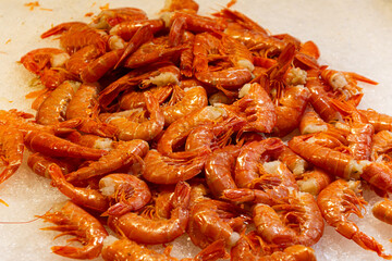 A large, appetizing pile of freshly cooked, orange-red large shrimp (gambas/prawns) displayed on a bed of crushed ice, ready for sale at a seafood market.

