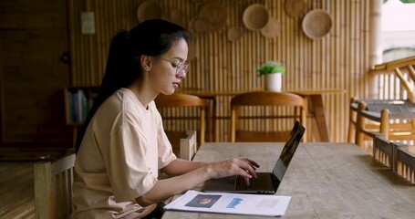 Asian woman adult sits at table working on laptop reviewing bar charts and pie graphs related to garden planning and organic farm progress using digital technology in natural home workspace - Powered by Adobe