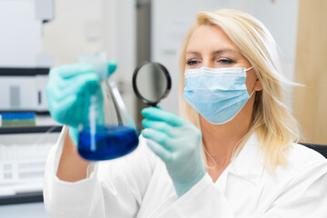 Middle aged female scientist wearing mask in lab, studying chemical solution in flask through magnifying glass.