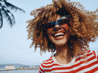 Vibrant close portrait of a joyful person with curly hair, bold sunglasses, and a striped shirt, captured under bright sun against a breezy seaside backdrop.