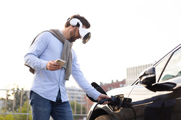 A man wearing VR glasses connects an electric vehicle to a charging station while holding a phone.