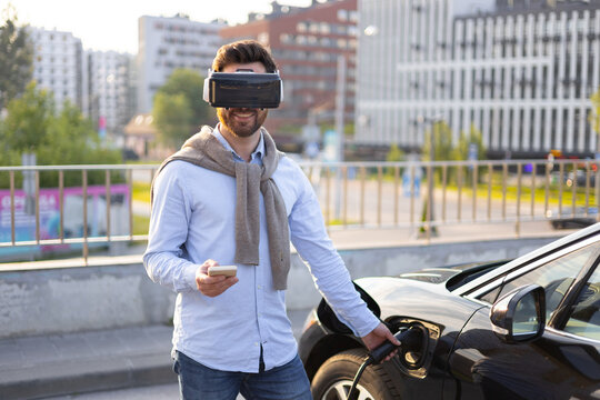 A man wearing a VR headset charges his electric vehicle while holding a smartphone, showcasing modern technology and sustainable transportation.