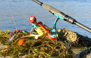 Carbon spinning rod, lying on dry grass near a lake or river with a colored reel and silicone baits. Catching walleye on a jig head and a pull-off leash. Fishing on the banks of a river or lake. 