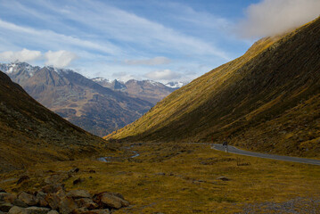 Golden slopes descend between rugged peaks. A winding road vanishes into the alpine distance. Snowy summits rise beyond the quiet valley.