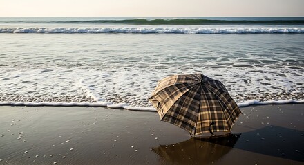 A plaid umbrella lies abandoned on a wet sandy beach with gentle ocean waves rolling in the background under a bright sky.