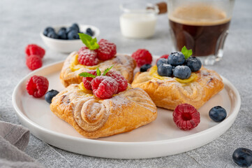 Danish puff pastry cakes with custard cream and fresh berries, sprinkled with powdered sugar on a white plate on a concrete background.