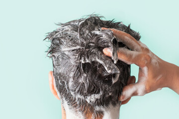 Male hands wash their hair with shampoo and foam on blue background close-up.