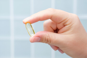 A hand holds a yellow fish oil capsule against a blue tile background, close-up.